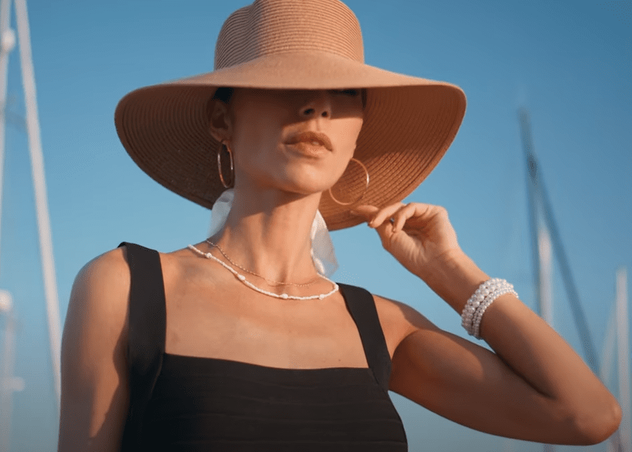 A woman wearing a wide-brimmed straw hat and black sleeveless dress stands outdoors. Her must-have accessories include hoop earrings, a necklace, and layered pearl bracelets, with the clear blue sky and sailboat masts in the background.